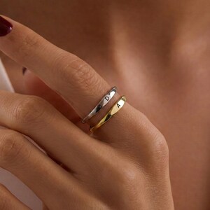 May include: Two rings, one silver and one gold, are worn on a finger. The silver ring has the letter "D" engraved on it, and the gold ring has the letter "A". The rings are simple bands with a slightly curved design.