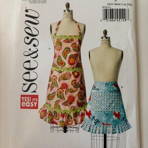 May include: A See & Sew B5943 sewing pattern envelope featuring two apron designs. Design A is a pink paisley apron with green rickrack trim and a ruffled hem. Design B is a blue apron with a red bird and floral print and a ruffled hem.