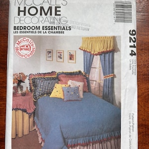 May include: A McCall's Home Decorating Bedroom Essentials sewing pattern envelope, number 9214. The front features an illustration of a bed with a blue bedspread, ruffled edges, and decorative pillows, alongside a matching curtain set and table cover.