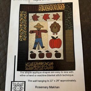 May include: A sewing pattern titled Signs of Autumn featuring a wall hanging design with a scarecrow, turkey, pumpkin, and apples. The pattern includes instructions for applique shapes and states the finished wall hanging is 22 inches by 28 inches.