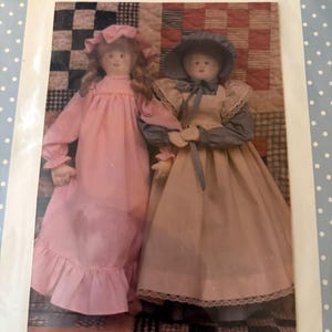 May include: Two fabric dolls with painted faces. The left doll wears a pink long-sleeved dress with a matching bonnet. The right doll wears a blue long-sleeved dress with a tan apron and a blue bonnet. Both dolls have yarn hair.