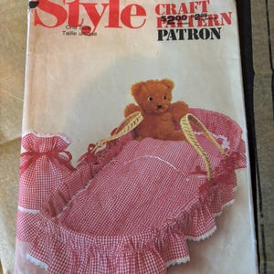 May include: A Style craft pattern envelope 3653 featuring a brown teddy bear sitting in a red and white gingham doll bassinet with a matching drawstring bag. The bassinet has a ruffled edge with white lace trim and woven handles.