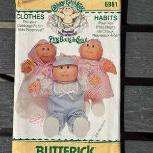 Puede incluir: Un sobre de patrón de costura Butterick para ropa de muñecas Cabbage Patch Kids Preemies, número 6981. El paquete presenta ilustraciones de tres muñecas con varios atuendos, incluyendo un vestido rosa con capucha, un pelele azul y un vestido rosa.