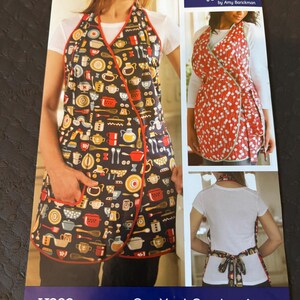May include: A sewing pattern for a One-Yard Overlap Apron by Indygo Junction, featuring a dark blue apron with a colourful kitchen utensil print and a red-orange apron with a white dandelion print, shown from the front and back views.