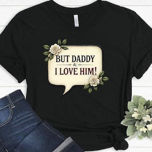Puede incluir: Camiseta negra con una burbuja de diálogo blanca que dice "But Daddy & I Love Him!" con flores blancas y hojas verdes alrededor de la burbuja de diálogo.