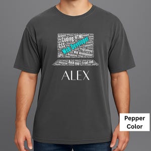 May include: A dark grey t-shirt featuring the name "ALEX" in white. Above the name is a laptop graphic composed of words such as "Coding", "HTML", and "Web Developer". The shirt is a pepper colour.