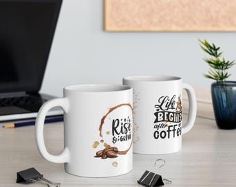May include: Two white ceramic coffee mugs with black text and coffee bean graphics. One mug says "Rise & Grind" inside a coffee stain circle. The other mug says "Life begins after coffee".