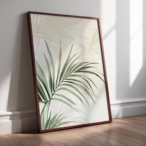 Puede incluir: Una impresión botánica enmarcada con una ilustración de acuarela de hojas de palma verdes sobre un fondo beige suave. La obra de arte está en un marco de madera marrón oscuro y se apoya contra una pared blanca.