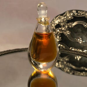 May include: A clear glass perfume bottle with a teardrop shape and a matching glass stopper. The bottle is filled with a dark amber liquid and sits upright, displaying a smooth, transparent surface and a rounded base.