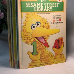 May include: A stack of vintage Sesame Street Library books. The cover of Volume 1 features Big Bird holding a green number 1 and the letters A and B. The title is in green and white, with the words "Featuring the Letters A and B and the Number 1."