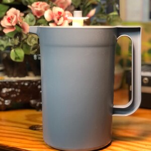 Può includere: Una brocca di plastica blu-grigio con manico e coperchio bianco. La brocca è cilindrica e poggia su una superficie di legno. Lo sfondo include una composizione floreale.