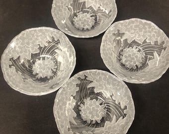 May include: Four clear glass bowls with a floral design. Each bowl features a central flower surrounded by swirling lines and a border of smaller flowers. The bowls are arranged on a dark surface.