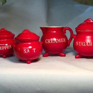 Puede incluir: Un juego de cuatro recipientes de cocina de cerámica roja. El juego incluye dos botes con tapa etiquetados "PEPPER" y "SALT", una jarra de crema etiquetada "CREAMER" y un azucarero con tapa etiquetado "SUGAR".