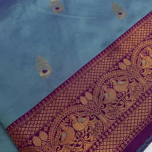 Op de afbeelding: Een close-up van een blauwgrijs textiel met een decoratieve rand. De rand is diep paars met goudkleurige patronen met vogels en geometrische ontwerpen. Het textiel heeft goudkleurige, bladachtige accenten.