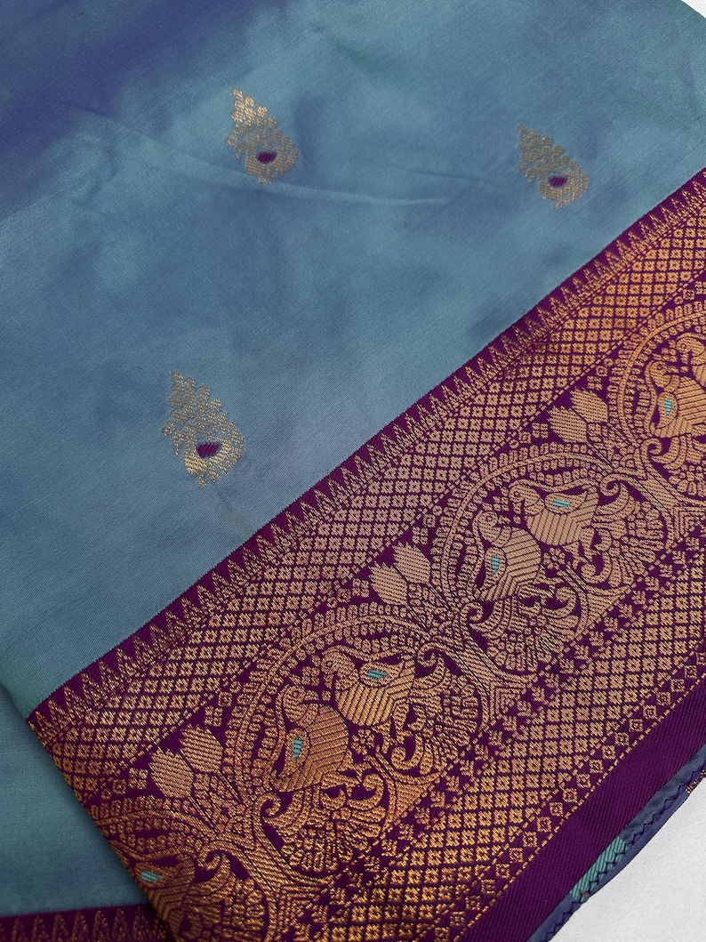 Op de afbeelding: Een close-up van een blauwgrijs textiel met een decoratieve rand. De rand is diep paars met goudkleurige patronen met vogels en geometrische ontwerpen. Het textiel heeft goudkleurige, bladachtige accenten.