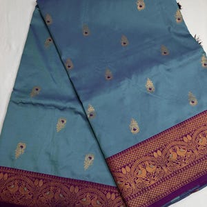 Op de afbeelding: Een turquoise textiel met een decoratieve rand en gouden accenten. De stof heeft een herhalend patroon van gouden druppelvormen en een gedetailleerde rand met gouden en paarse ontwerpen. Het textiel is gevouwen en toont de textuur en kleurvariaties.