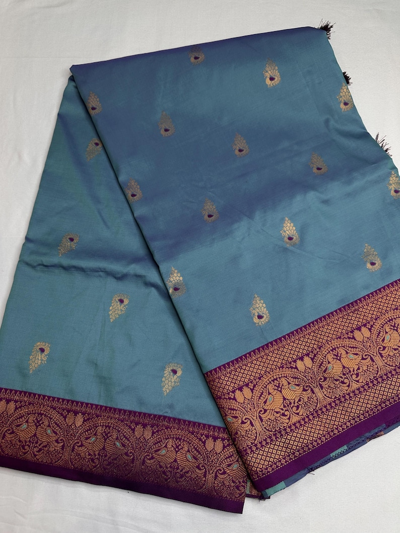 Op de afbeelding: Een turquoise textiel met een decoratieve rand en gouden accenten. De stof heeft een herhalend patroon van gouden druppelvormen en een gedetailleerde rand met gouden en paarse ontwerpen. Het textiel is gevouwen en toont de textuur en kleurvariaties.