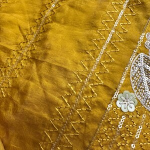May include: Mustard-colored fabric with intricate gold zigzag embroidery and delicate silver sequins. The design includes small floral embellishments and a subtle sheen, creating a luxurious texture. The fabric appears to be silk or a similar material.