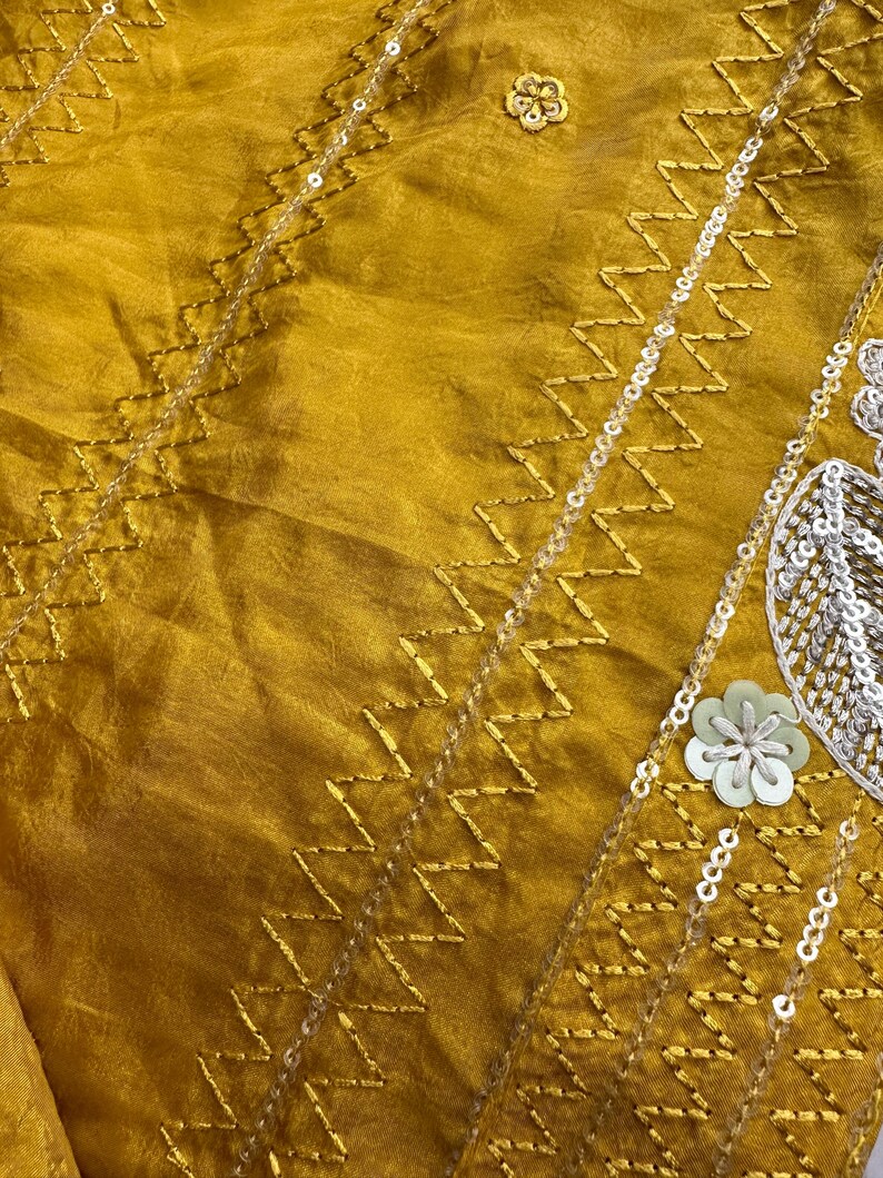 May include: Mustard-colored fabric with intricate gold zigzag embroidery and delicate silver sequins. The design includes small floral embellishments and a subtle sheen, creating a luxurious texture. The fabric appears to be silk or a similar material.