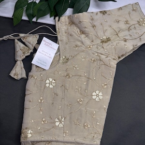 May include: A beige blouse with intricate floral embroidery and sequin embellishments. The blouse features short sleeves and a round neckline. A tag reads "Sheetal's FabFashion Jewelry & Sarees Boutique Your Style, Our Passion."