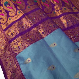 Op de afbeelding: Een luxe zijden sari met een levendig paars en goud paisley design. De ingewikkelde rand heeft gedetailleerde patronen en het onderste gedeelte is zacht blauwgrijs met gouden accenten. De stof is gevouwen en toont zijn rijke textuur en kleur.