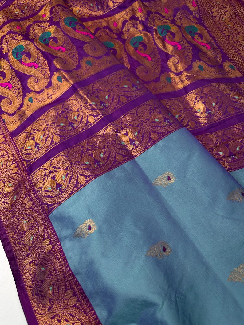 Op de afbeelding: Een luxe zijden sari met een levendig paars en goud paisley design. De ingewikkelde rand heeft gedetailleerde patronen en het onderste gedeelte is zacht blauwgrijs met gouden accenten. De stof is gevouwen en toont zijn rijke textuur en kleur.