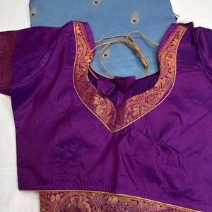 Op de afbeelding: Een paarse blouse met een diepe V-hals en gouden bies. De blouse wordt gecombineerd met een lichtblauwe stof met geborduurde details en kwastjes. De blouse heeft een trekkoordsluiting.