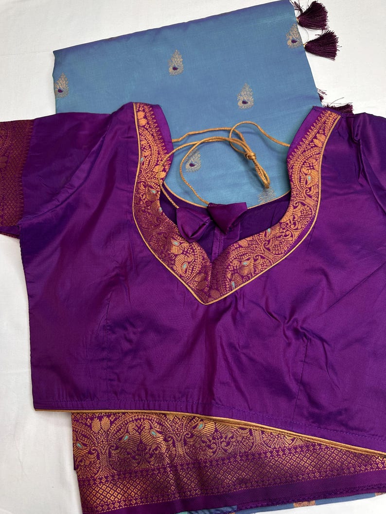 Op de afbeelding: Een paarse blouse met een diepe V-hals en gouden bies. De blouse wordt gecombineerd met een lichtblauwe stof met geborduurde details en kwastjes. De blouse heeft een trekkoordsluiting.