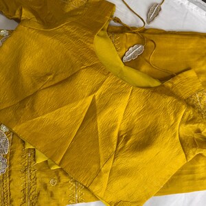 May include: Mustard yellow silk blouse and matching fabric, with intricate embroidery and embellishments. A delicate necklace with leaf-shaped pendants rests nearby. The blouse features a textured, pleated design.