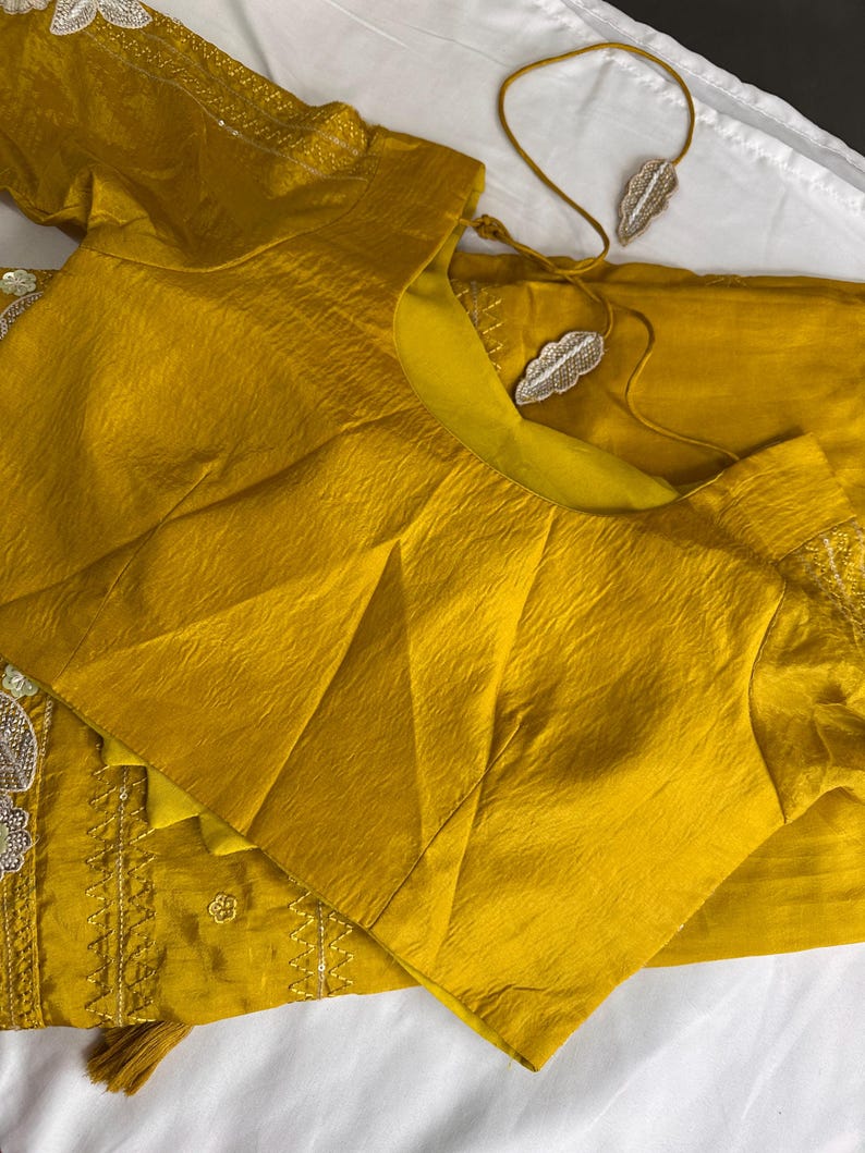 May include: Mustard yellow silk blouse and matching fabric, with intricate embroidery and embellishments. A delicate necklace with leaf-shaped pendants rests nearby. The blouse features a textured, pleated design.