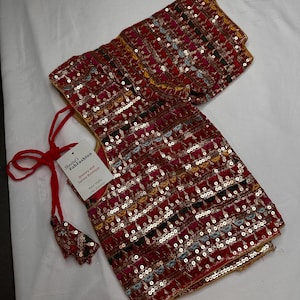May include: A red, sequined blouse with gold trim and a matching tassel. The blouse features intricate embroidery and a tag that reads "Sheetal's FabFashion Jewelry and Sarees Boutique". The garment is displayed on a white surface.