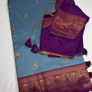 Op de afbeelding: Een blauwgrijze sari met gouden geborduurde accenten en een decoratieve rand. De sari wordt gecombineerd met een paarse blouse met gouden randen en bijpassende borduursels. Kwasten en trekkoorden voegen details toe aan de blouse.