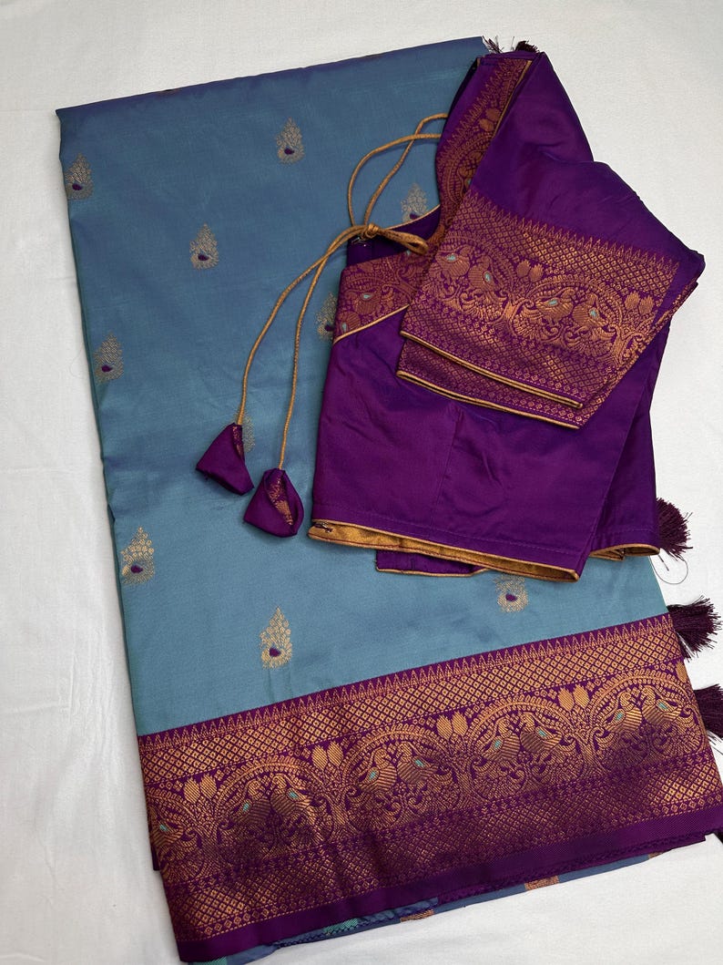 Op de afbeelding: Een blauwgrijze sari met gouden geborduurde accenten en een decoratieve rand. De sari wordt gecombineerd met een paarse blouse met gouden randen en bijpassende borduursels. Kwasten en trekkoorden voegen details toe aan de blouse.