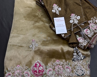May include: A gold-toned saree with intricate embroidery in pink, silver, and green. The border features detailed floral patterns and embellishments. A matching brown blouse with similar embroidery and a tag that reads "Sheetal's TabFashion Jewelry & Sarees Boutique" is included.