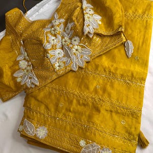 May include: Mustard yellow saree and blouse set. The blouse features intricate floral embroidery with silver and white accents. The saree has a matching border with similar embellishments and tassels. The fabric appears to be a lightweight, flowing material.