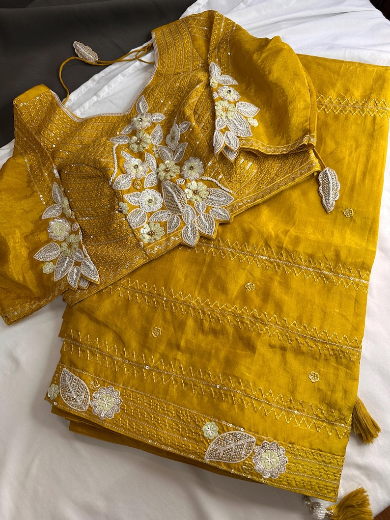 May include: Mustard yellow saree and blouse set. The blouse features intricate floral embroidery with silver and white accents. The saree has a matching border with similar embellishments and tassels. The fabric appears to be a lightweight, flowing material.