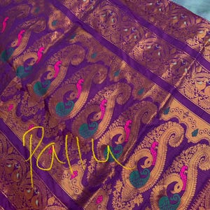 Op de afbeelding: Close-up van een paars textiel met gouden paisley patronen en pauw motieven. Het ontwerp bevat roze en groene accenten. Het woord "Pallu" is in het geel over de stof geschreven. Het textiel heeft een luxueuze uitstraling.