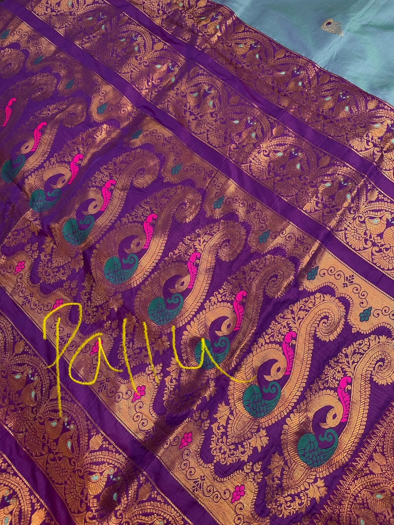Op de afbeelding: Close-up van een paars textiel met gouden paisley patronen en pauw motieven. Het ontwerp bevat roze en groene accenten. Het woord "Pallu" is in het geel over de stof geschreven. Het textiel heeft een luxueuze uitstraling.