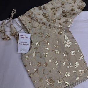 May include: A beige blouse featuring short sleeves and an intricate floral pattern embroidered with gold sequins. The garment includes a tie-back closure with decorative tassels and a white tag labeled Sheetals FabFashion Jewelry & Sarees Boutique.