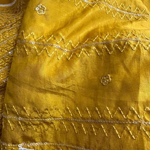 May include: Close-up of a mustard yellow fabric with intricate embroidery. The fabric features zigzag stitching with silver sequins and small floral embellishments. The texture appears soft and flowing, suggesting a garment or textile.