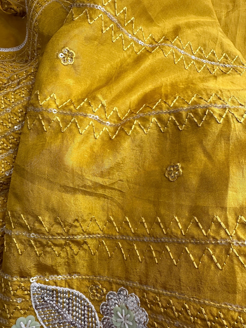 May include: Close-up of a mustard yellow fabric with intricate embroidery. The fabric features zigzag stitching with silver sequins and small floral embellishments. The texture appears soft and flowing, suggesting a garment or textile.
