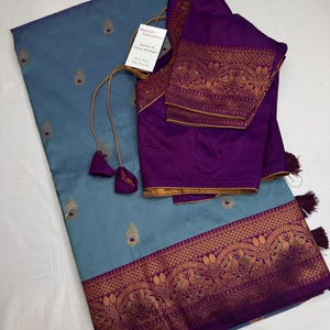 Op de afbeelding: Een blauwgrijze sari met gouden borduursel en een paarse rand. Een bijpassende paarse blouse met gouden afwerking en borduursel is inbegrepen. De sari heeft een label met "Sheetal's FabFashion Jewelry & Saree Boutique".