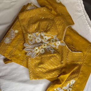 May include: Mustard yellow embroidered blouse and matching fabric. The blouse features intricate floral embroidery with white and silver accents. The fabric appears to be silk or a similar material, with a subtle sheen. The design is likely for a traditional garment.
