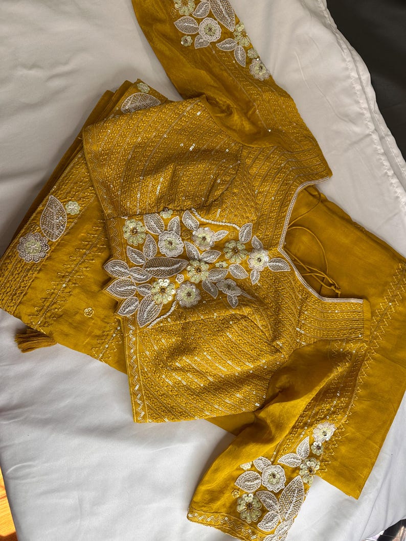 May include: Mustard yellow embroidered blouse and matching fabric. The blouse features intricate floral embroidery with white and silver accents. The fabric appears to be silk or a similar material, with a subtle sheen. The design is likely for a traditional garment.