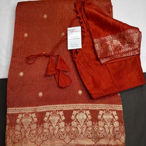 May include: A rust-colored saree featuring a subtle tonal pattern and small embroidered dots, paired with a matching blouse. The saree has a wide border with intricate gold floral motifs and a tag labeled Sheetal's FabFashion Jewelry & Sarees Boutique.