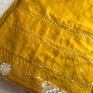 May include: Mustard yellow fabric with intricate gold embroidery and sequin embellishments. The design features zigzag stitching and floral and leaf motifs, creating a textured and decorative surface. The fabric has a slight sheen.