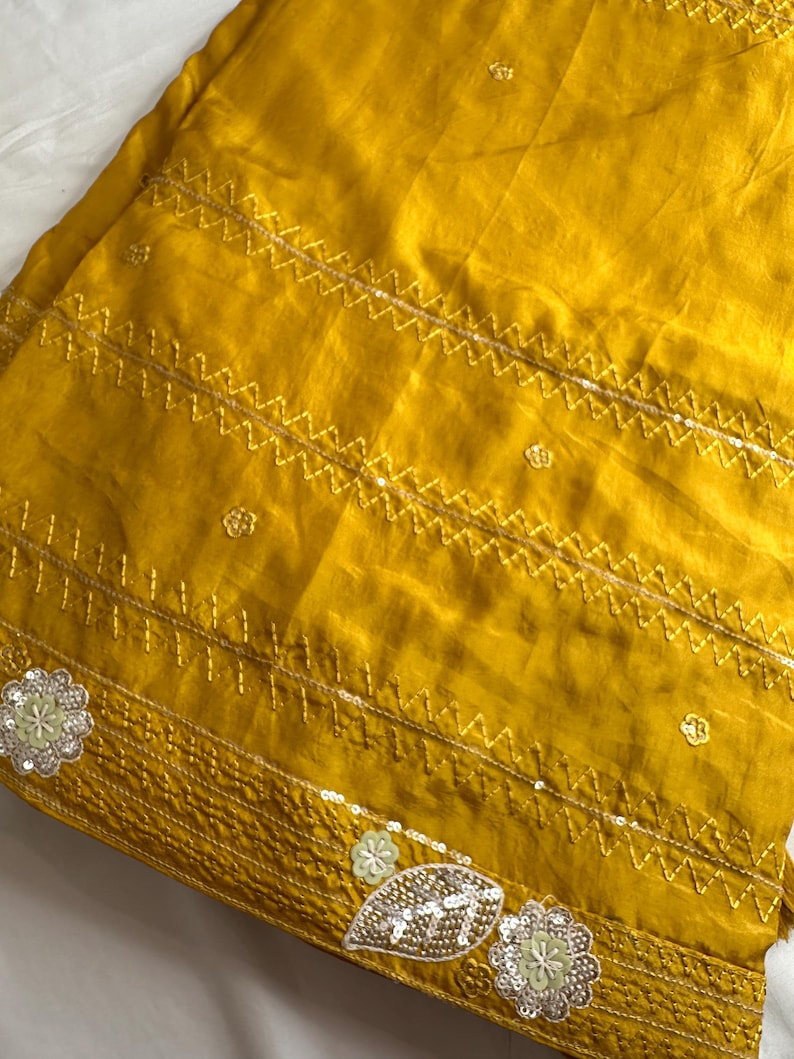 May include: Mustard yellow fabric with intricate gold embroidery and sequin embellishments. The design features zigzag stitching and floral and leaf motifs, creating a textured and decorative surface. The fabric has a slight sheen.
