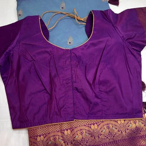 Op de afbeelding: Een levendige paarse blouse met een ronde hals en korte mouwen. Het wordt gecombineerd met een lichtblauwe stof met gouden borduursels en kwastjes. De onderrand van de stof heeft een sierlijk goudkleurig patroon.