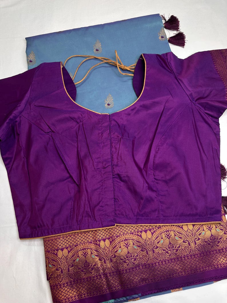 Op de afbeelding: Een levendige paarse blouse met een ronde hals en korte mouwen. Het wordt gecombineerd met een lichtblauwe stof met gouden borduursels en kwastjes. De onderrand van de stof heeft een sierlijk goudkleurig patroon.
