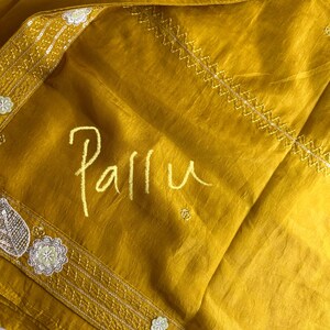 May include: Mustard yellow fabric with the word "Pallu" in a handwritten style. The fabric has decorative borders with floral and leaf designs, and a stitched pattern. The fabric is folded and appears to be silk or a similar material.